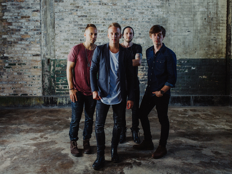 Four young men stand against a brick wall