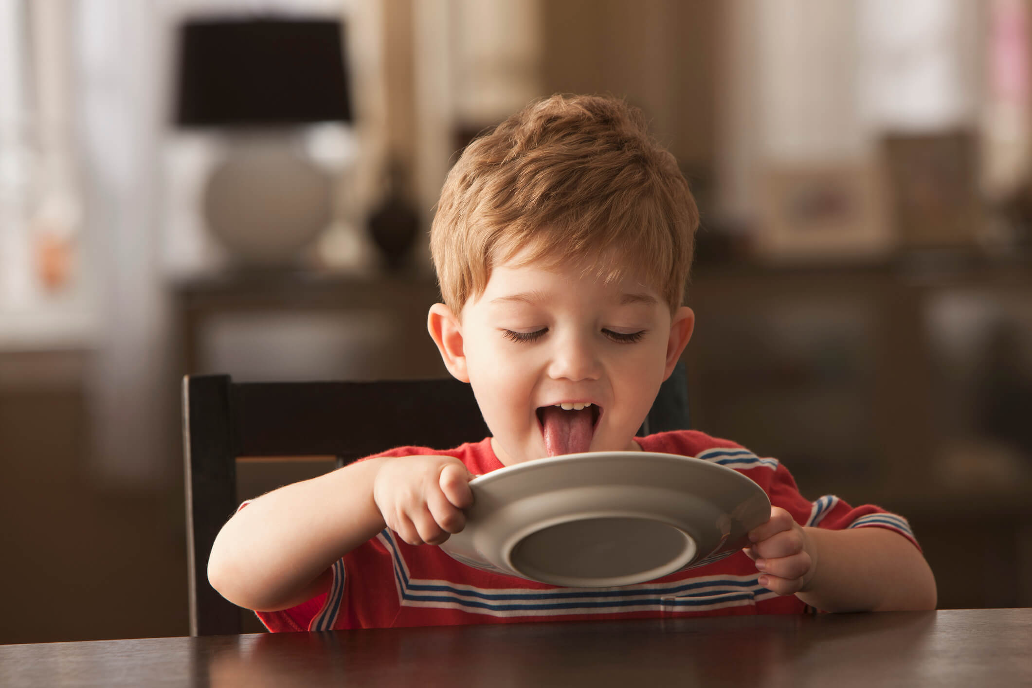 A little boy sitting at the table and licking his plate clean